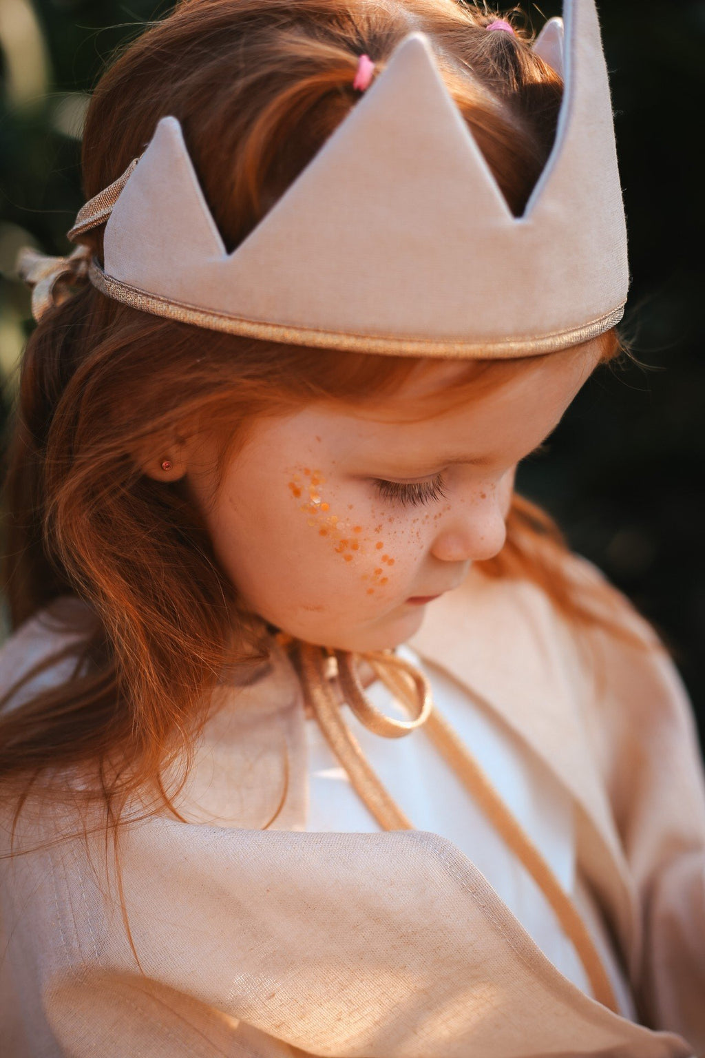 1. Young girl with red hair wearing a beige crown and cape, adorned with gold star dust glitter on her cheek, in an outdoor setting