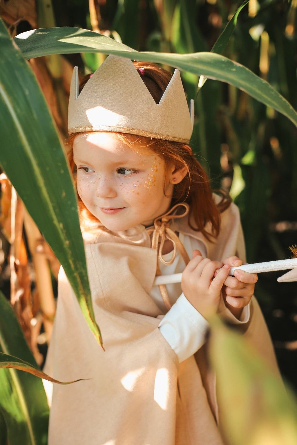 4. Young girl with red hair wearing a beige crown and cape, with gold star dust glitter on her cheek, peeking through green leaves