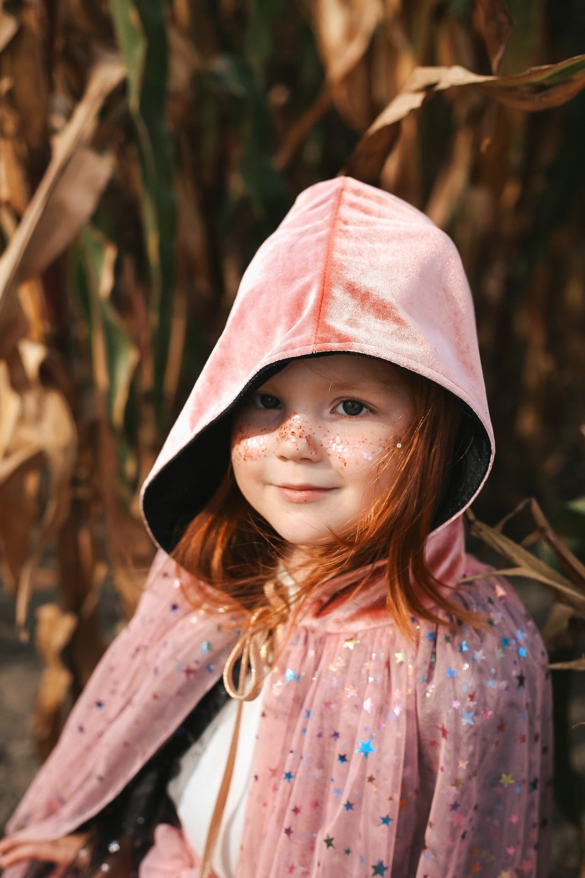 1. Young girl wearing pink hooded cape with decorative eco glitter on cheeks in outdoor setting