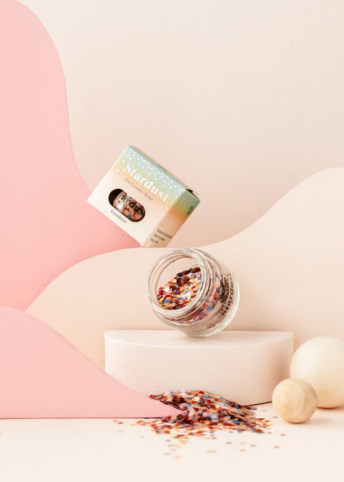 1. Glass jar of moi mili Rainbow Star Dust eco glitter with colorful confetti spilling out, set against a pastel pink and cream abstract background
