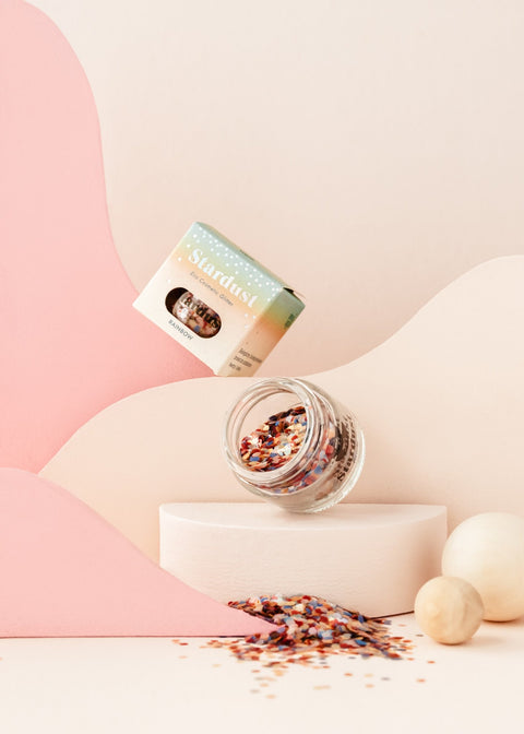 1. Glass jar of moi mili Rainbow Star Dust eco glitter with colorful confetti spilling out, set against a pastel pink and cream abstract background