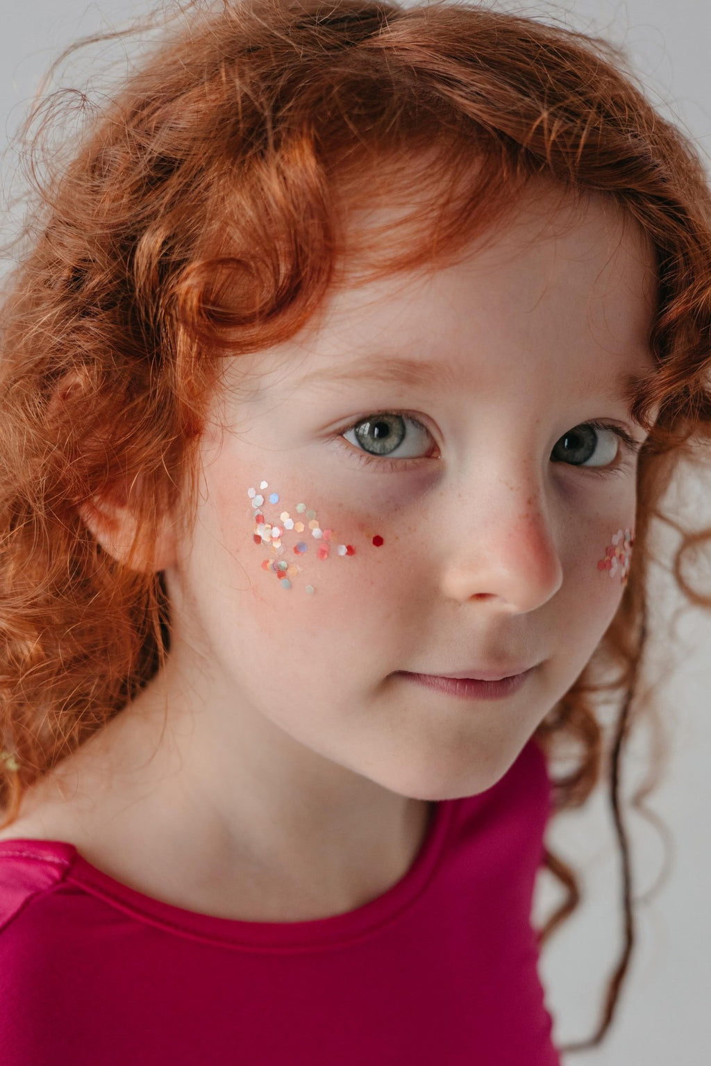 3. Close-up of a young girl with red hair wearing pink top, adorned with moi mili Rainbow Star Dust glitter on her cheeks