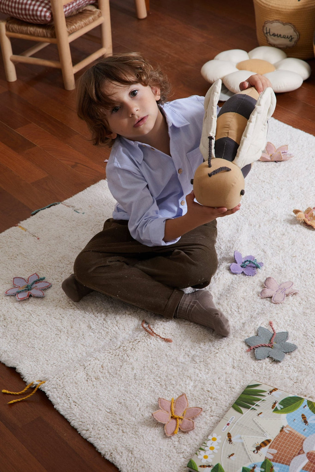 1. Child holding honey-colored bee-shaped pillow with dark gray stripes on white rug