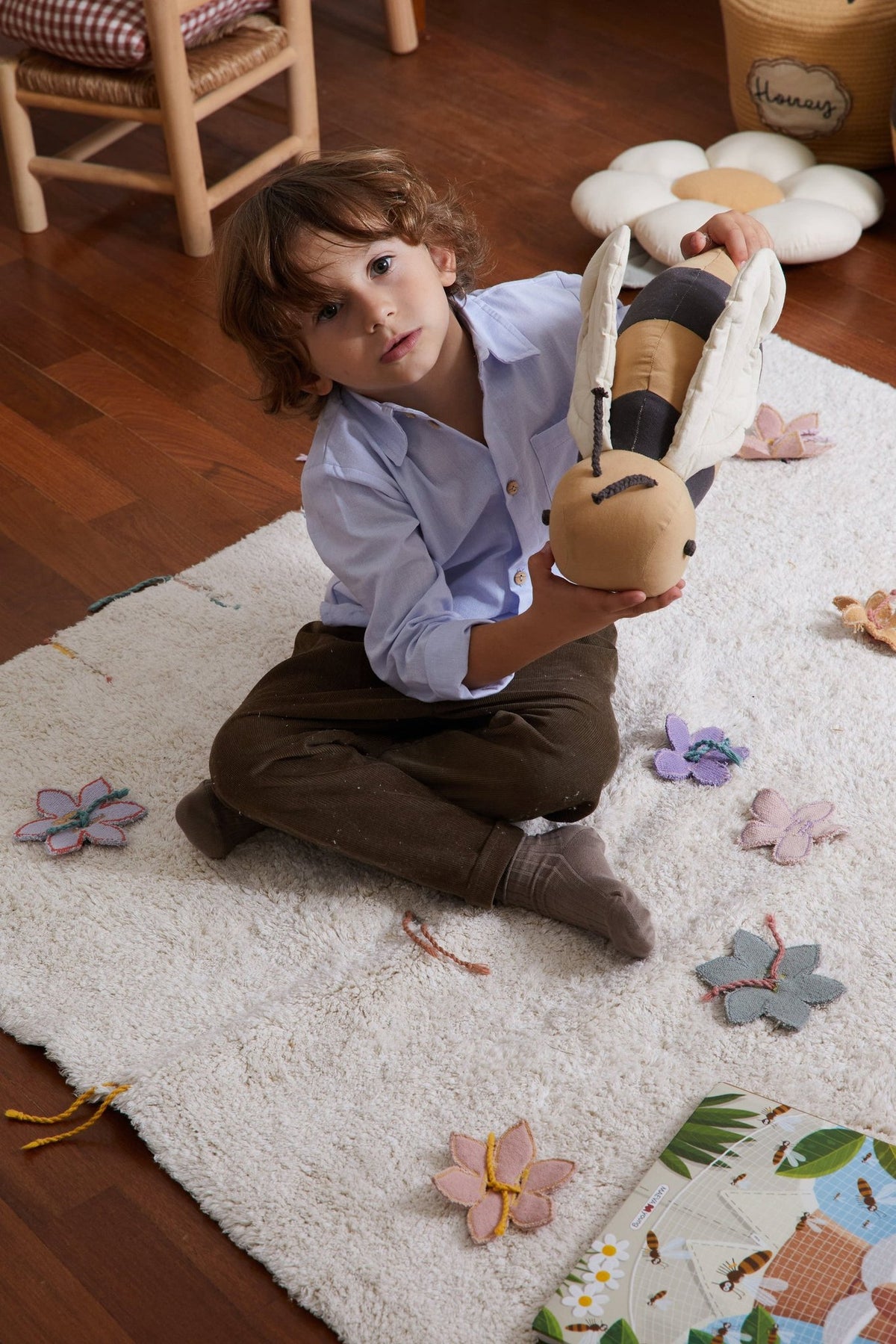 1. Child holding honey-colored bee-shaped pillow with dark gray stripes on white rug