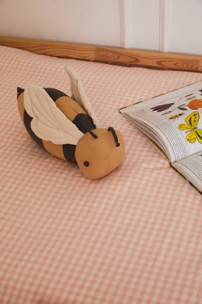 1. Honey-colored bee-shaped pillow with dark gray stripes on pink checkered bedspread next to open book