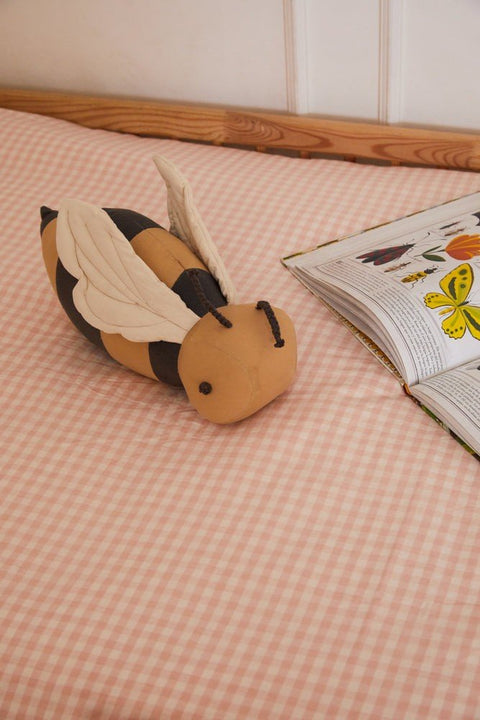 1. Honey-colored bee-shaped pillow with dark gray stripes on pink checkered bedspread next to open book