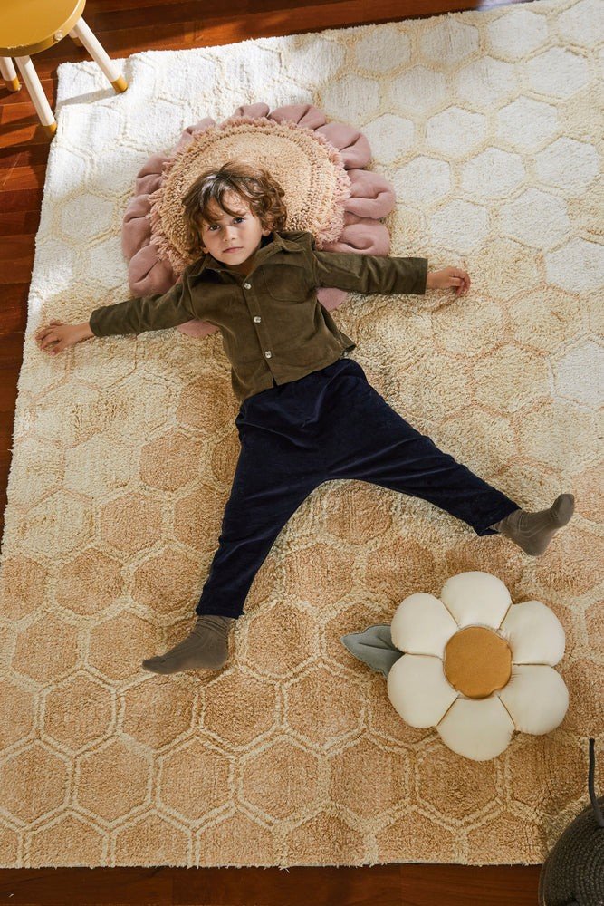 2. Child lying on a hexagonal patterned rug with a decorative daisy pillow nearby, featuring natural-colored petals and a honey center