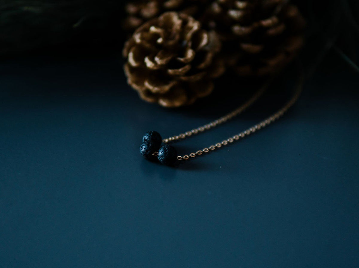7. Gold diffuser necklace with three black lava beads next to pine cones, lifestyle setting
