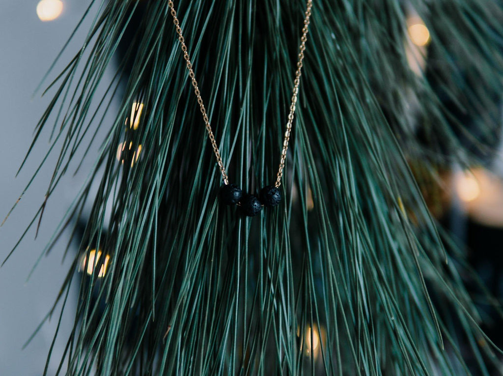 2. Gold diffuser necklace with three black lava beads hanging on a pine branch, lifestyle setting
