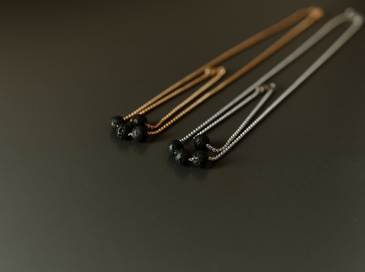 2. Black lava stone necklaces with three beads each, displayed on golden and silver stainless steel chains in a studio setting