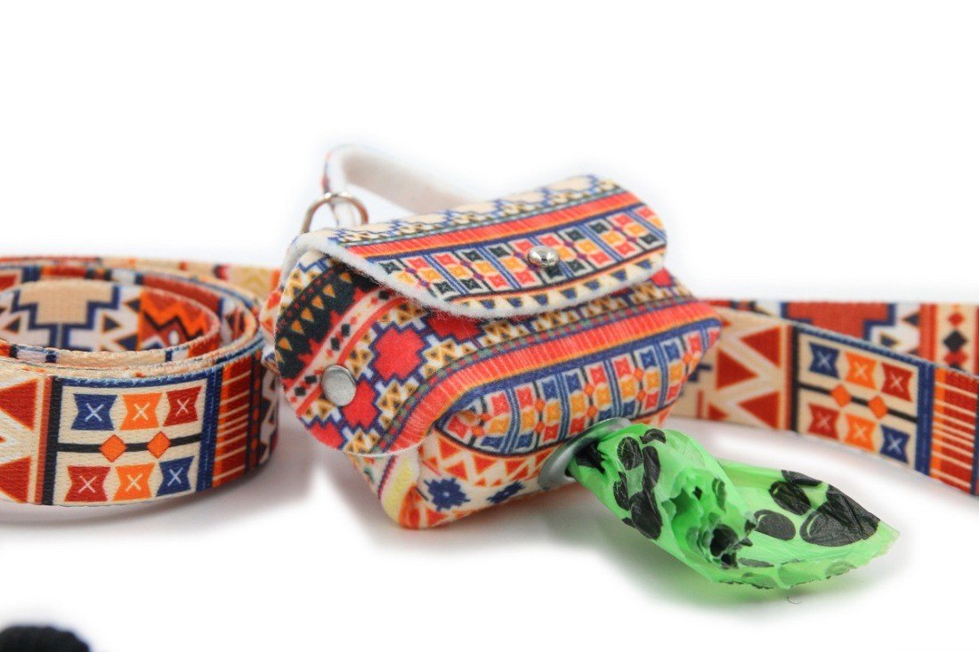 1. Boho-style dog waste bag dispenser with colorful geometric pattern attached to matching leash, green bag partially pulled out through metal eyelet