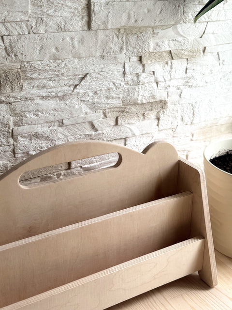 2. Montessori double-sided wooden book display on a wooden floor with a stone wall background, showcasing empty shelves and rounded handles