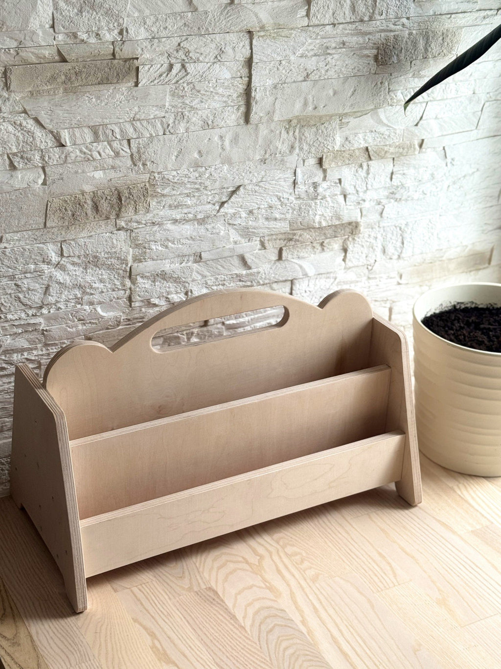 2. Montessori double-sided wooden book display on a wooden floor with a stone wall background, showcasing empty shelves and rounded handles
