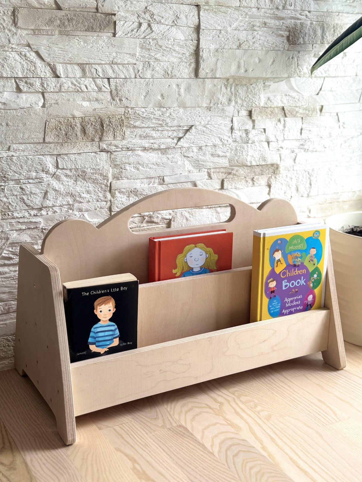 1. Montessori double-sided wooden book display with children's books on a wooden floor against a stone wall, featuring rounded handles and natural birch finish
