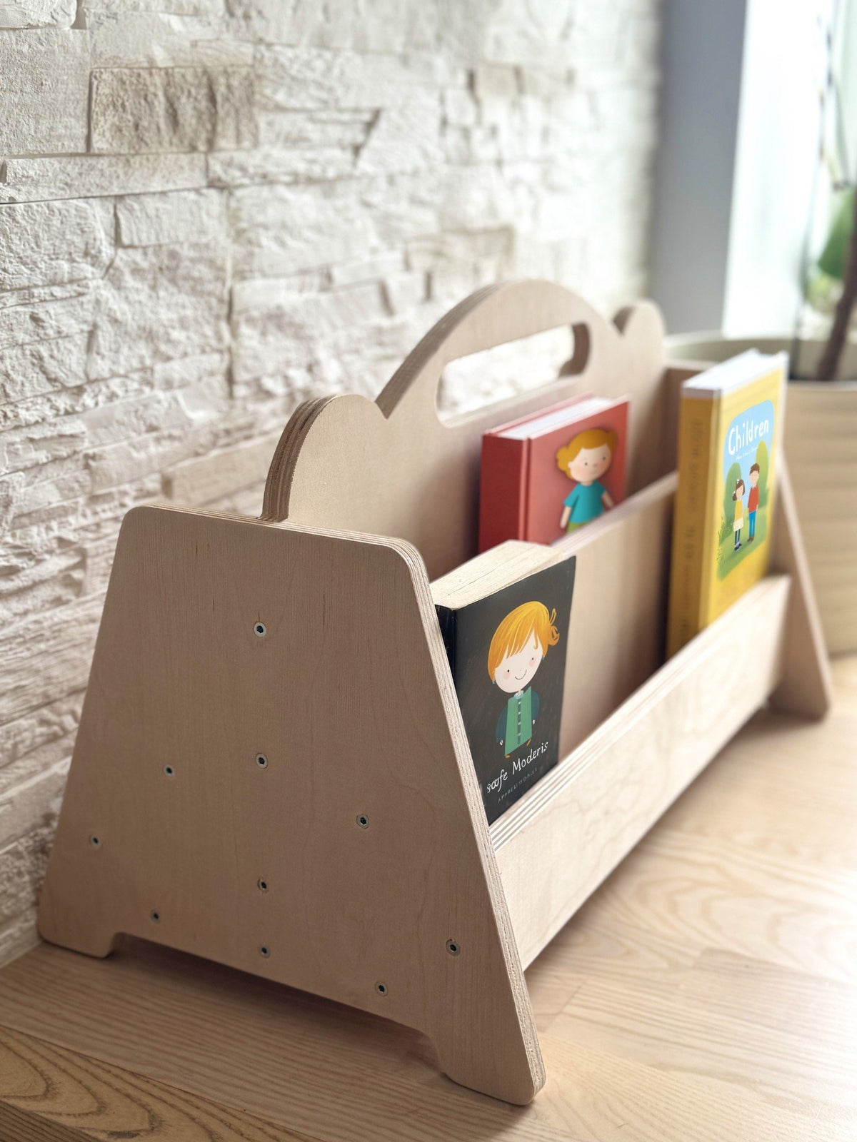 1. Montessori double-sided wooden book display with books on a wooden floor against a stone wall, featuring rounded handles and natural birch finish