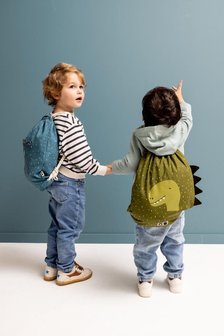 1. Two children wearing Trixie Baby drawstring bags, one green with dinosaur print, in a playful setting