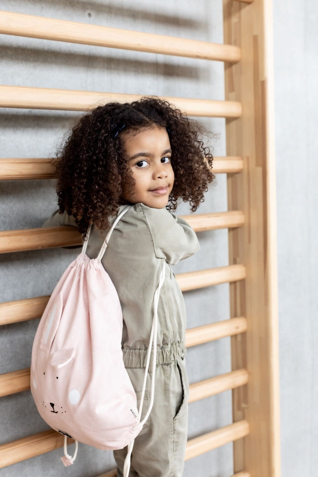 1. Child wearing pink rabbit-themed drawstring bag, standing by wooden bars
