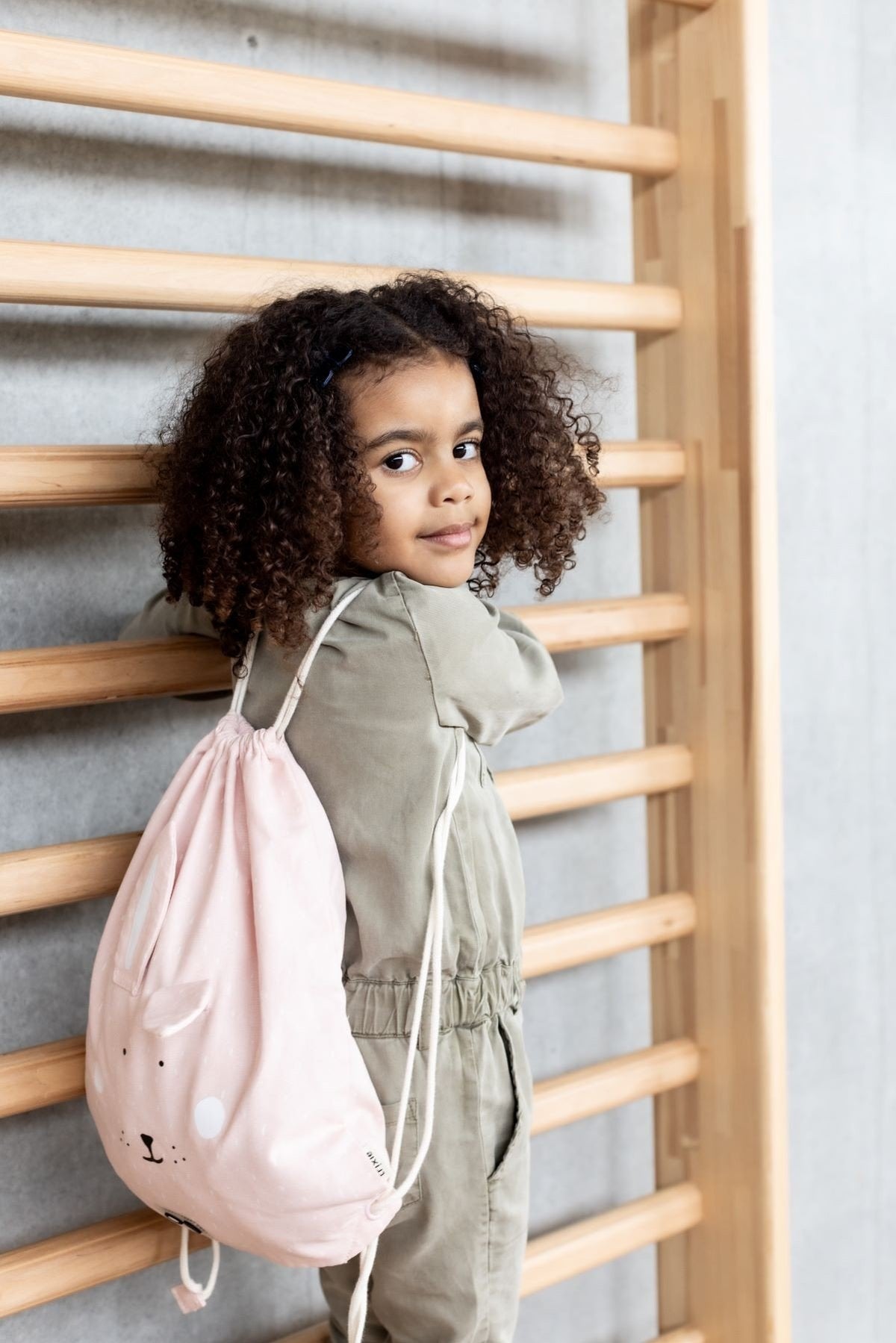 1. Child wearing pink rabbit-themed drawstring bag, standing by wooden bars
