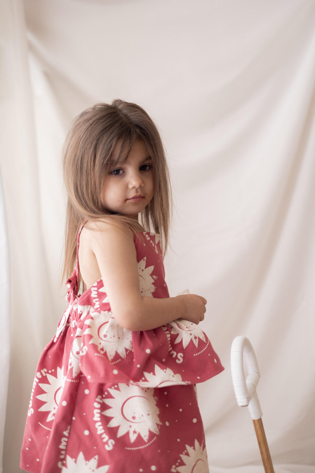 1. Girl wearing pink dress with sun prints standing in front of a white curtain