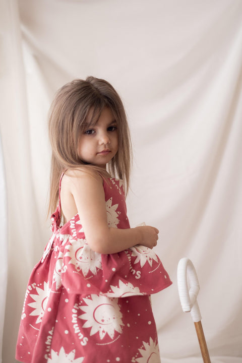 1. Girl wearing pink dress with sun prints standing in front of a white curtain