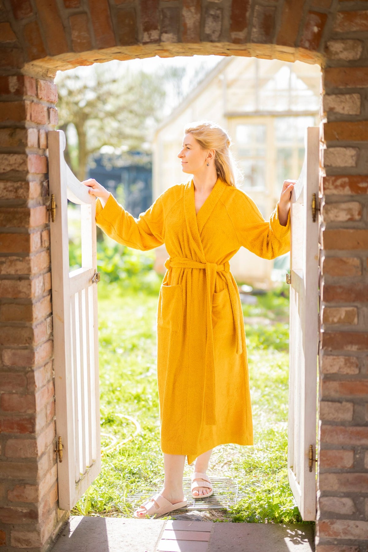 2. Woman wearing a yellow bamboo terry bathrobe by RÄTT, standing in a garden setting, highlighting the robe's shawl collar and belt closure
