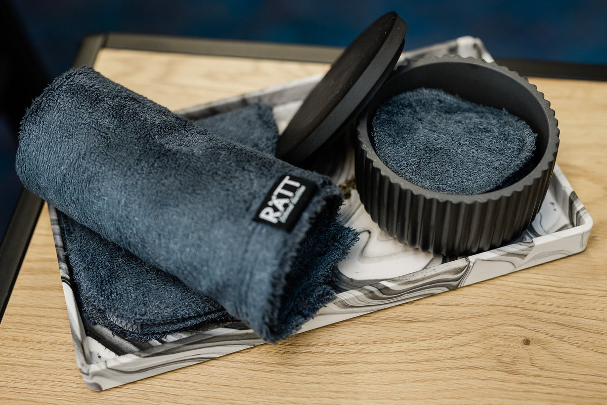 3. Close-up of RÄTT Home Spa Set with black jesmonite container, dark gray bamboo terry towels, and cleansing pads on a marbled tray, placed on a wooden surface