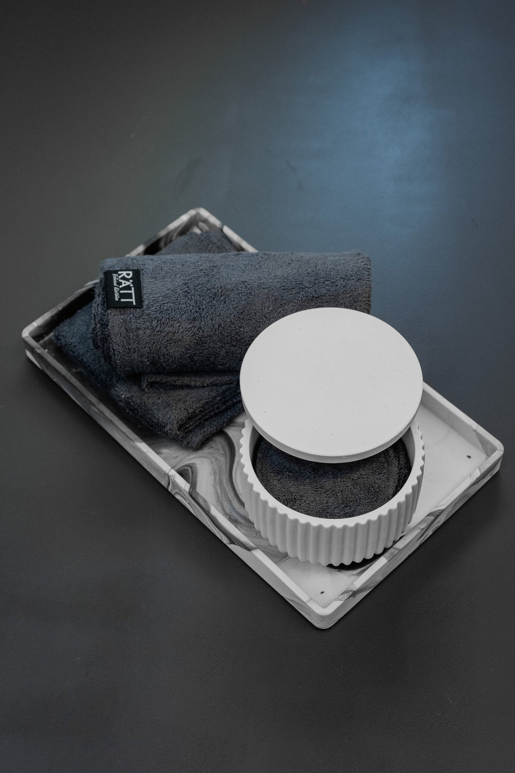 1. RÄTT Home Spa Set with white jesmonite container, dark gray bamboo terry towels, and cleansing pads on a marbled tray, displayed on a dark surface