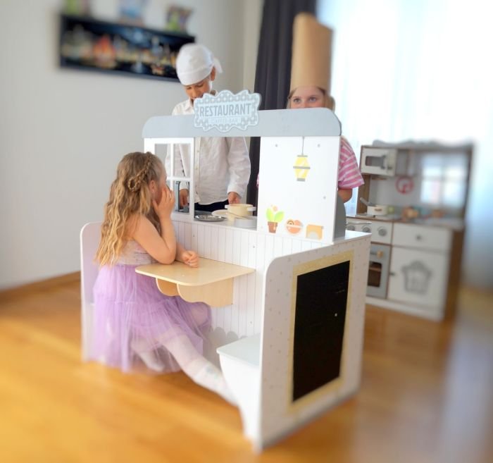 1. Three children playing with wooden 2in1 diner, one seated at table, others in chef hats in a playroom setting