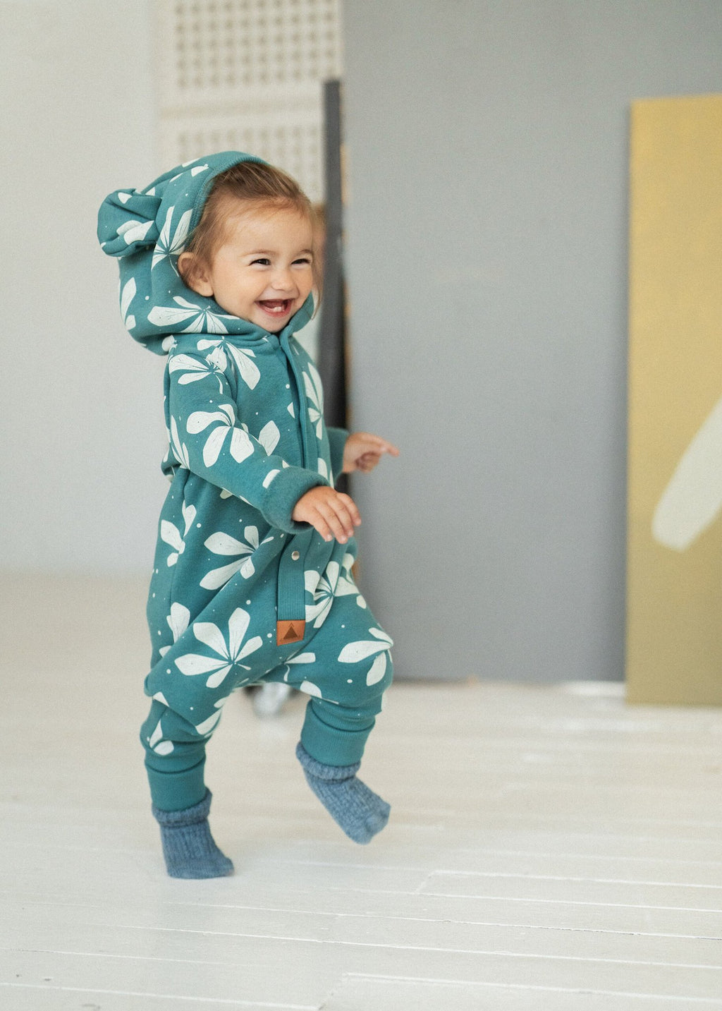 1. Toddler wearing green jumpsuit with chestnut leaf print and hood with ears, standing on white wooden floor