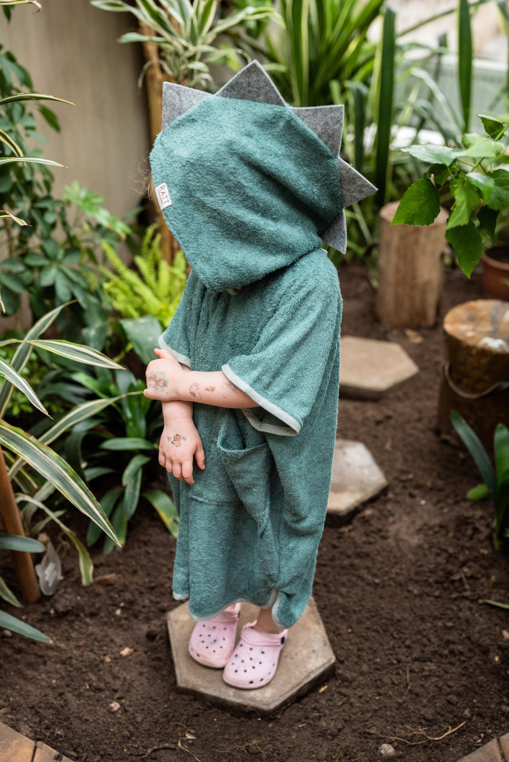 2. Child wearing RÄTT green dinosaur poncho with hood, side view in garden