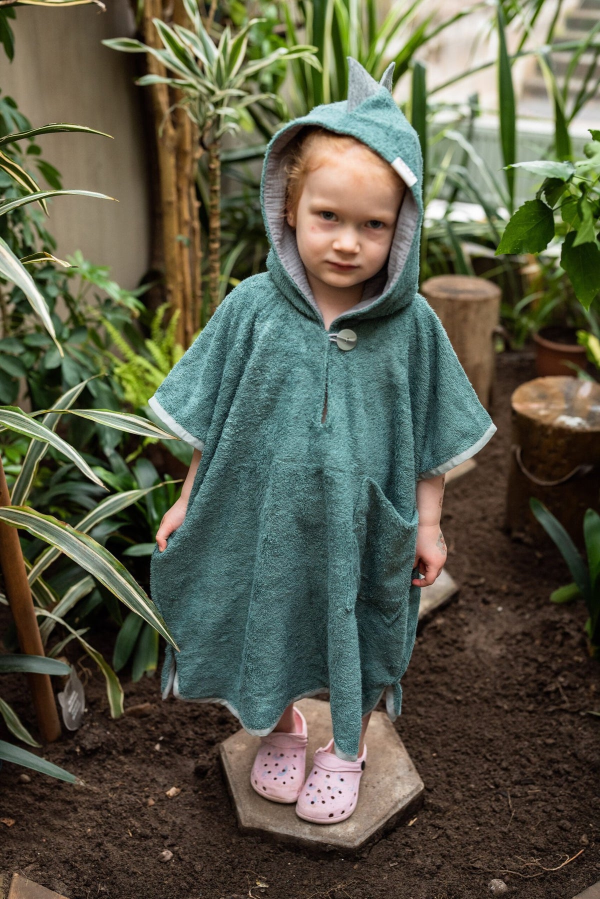 3. Front view of child in RÄTT green dinosaur poncho with hood, standing in garden
