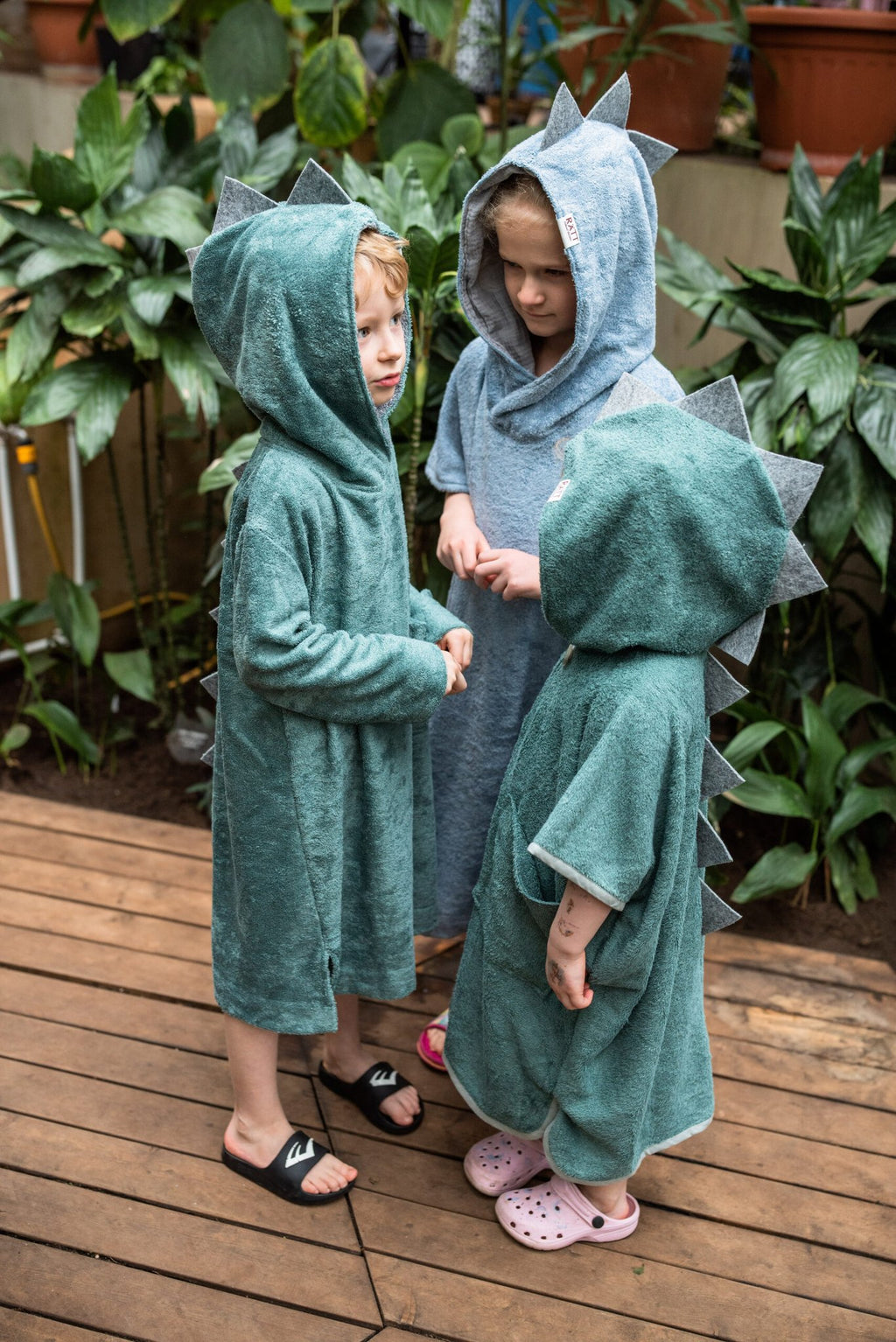 1. Toddlers wearing RÄTT short-sleeve dinosaur ponchos in green and blue, shown in a garden setting