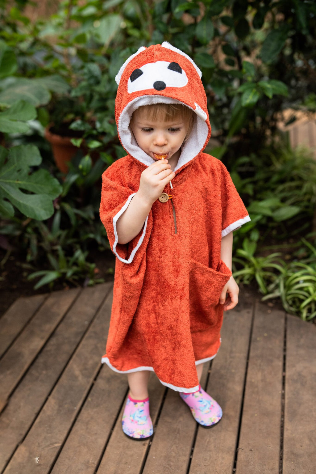 2. Toddler in RÄTT Short-Sleeve Poncho FOX, standing on wooden deck with greenery