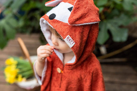 1. Toddler wearing RÄTT Short-Sleeve Poncho FOX with hood, shown outdoors with flowers