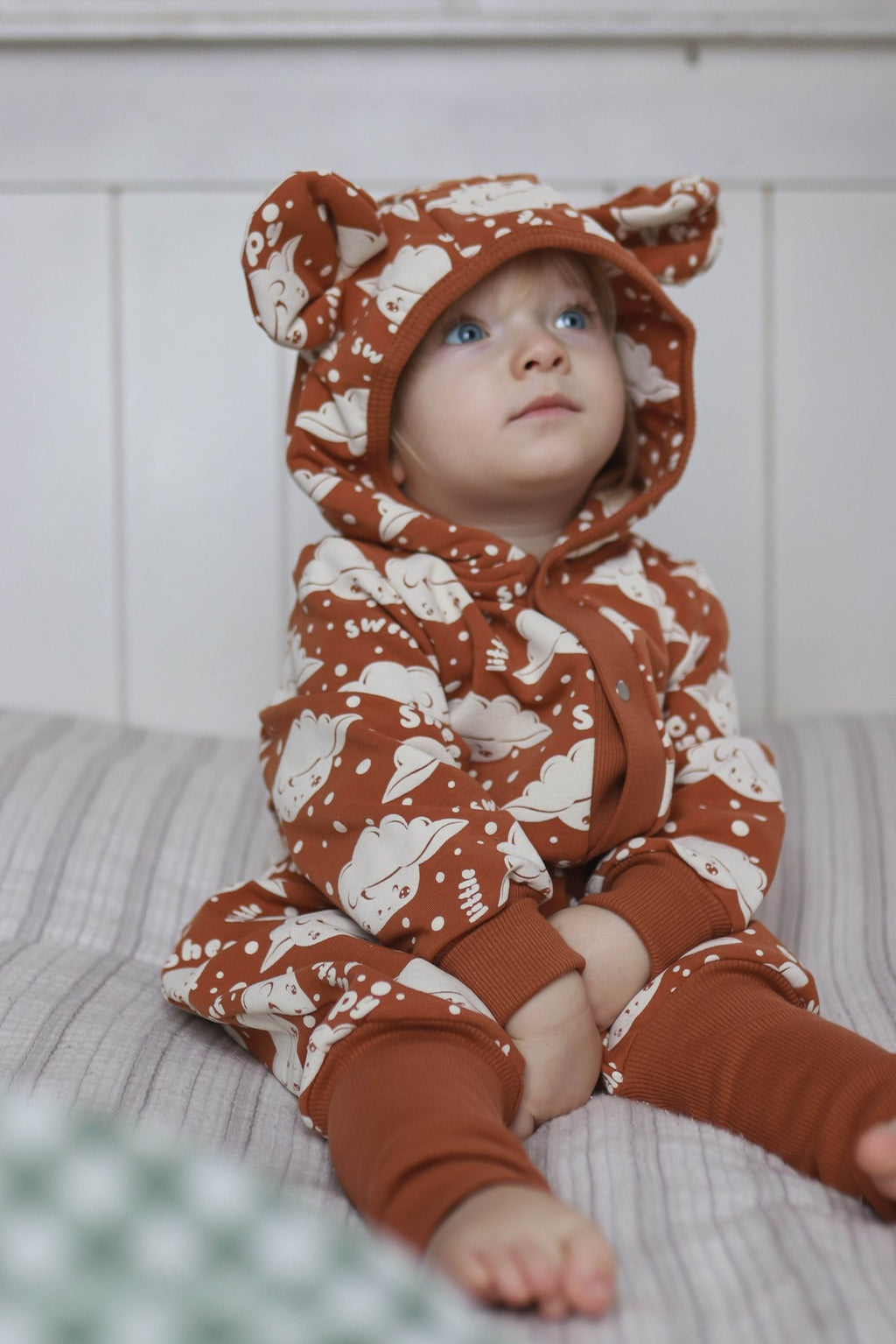 1. Baby wearing cinnamon-colored jumpsuit with sheep print and hood with ears, sitting on a bed