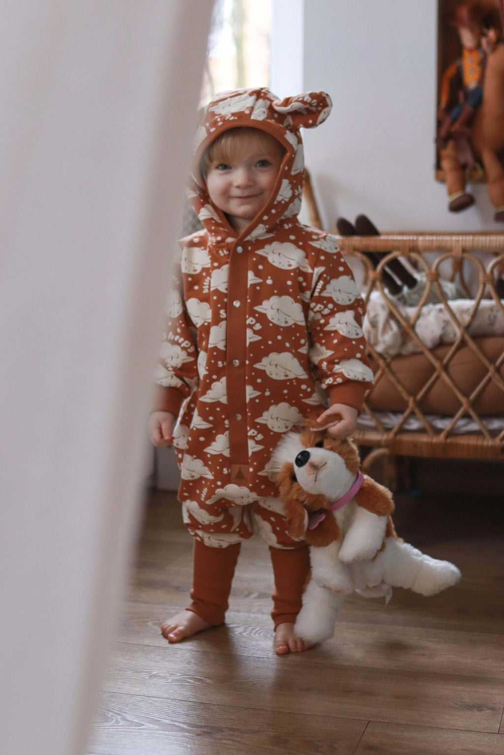2. Child standing in cinnamon sheep print jumpsuit with hood and ears, holding a stuffed toy in a cozy room