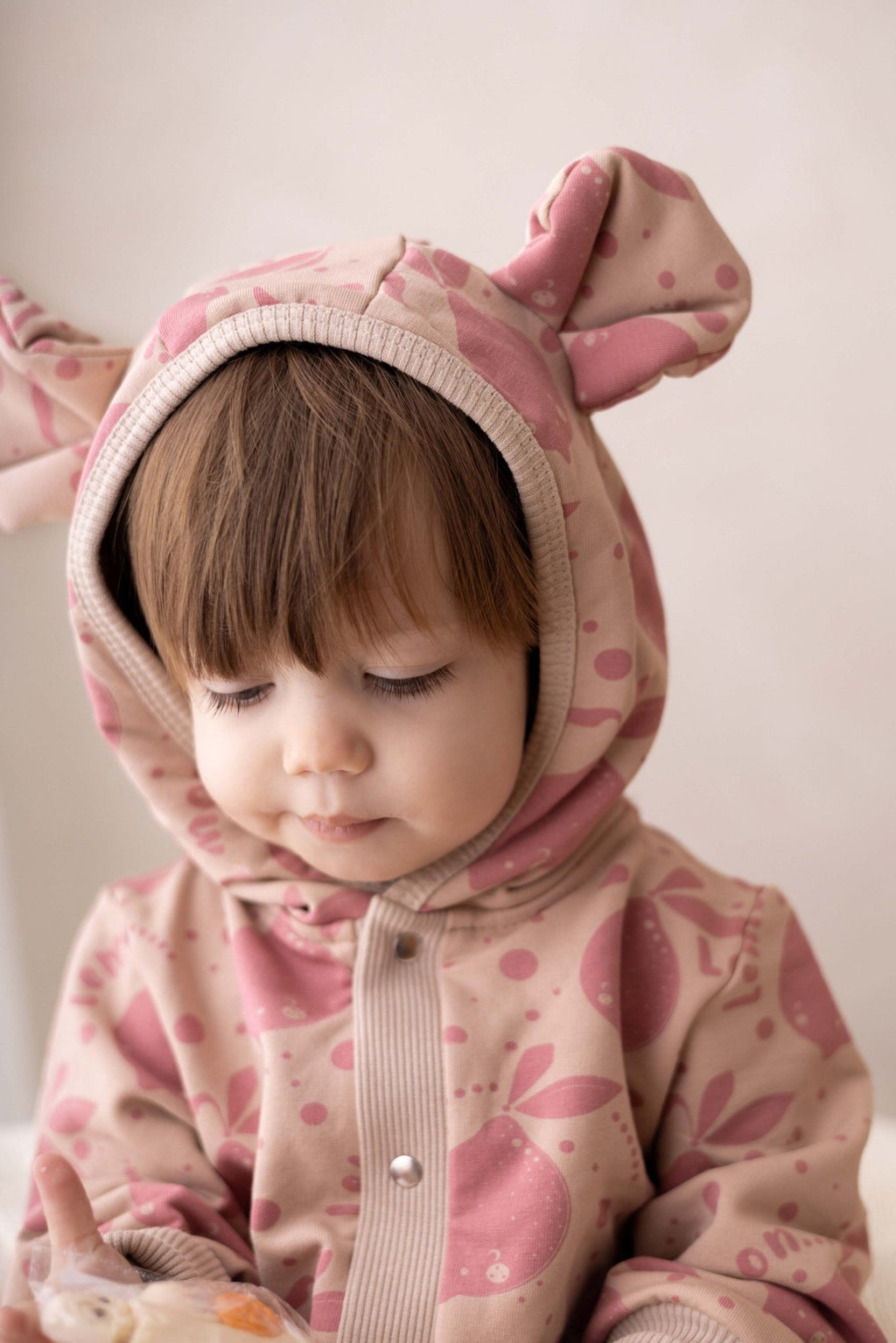 1. Baby looking down wearing Zezuzulla beige jumpsuit with pink lemon print and hood with ears indoors