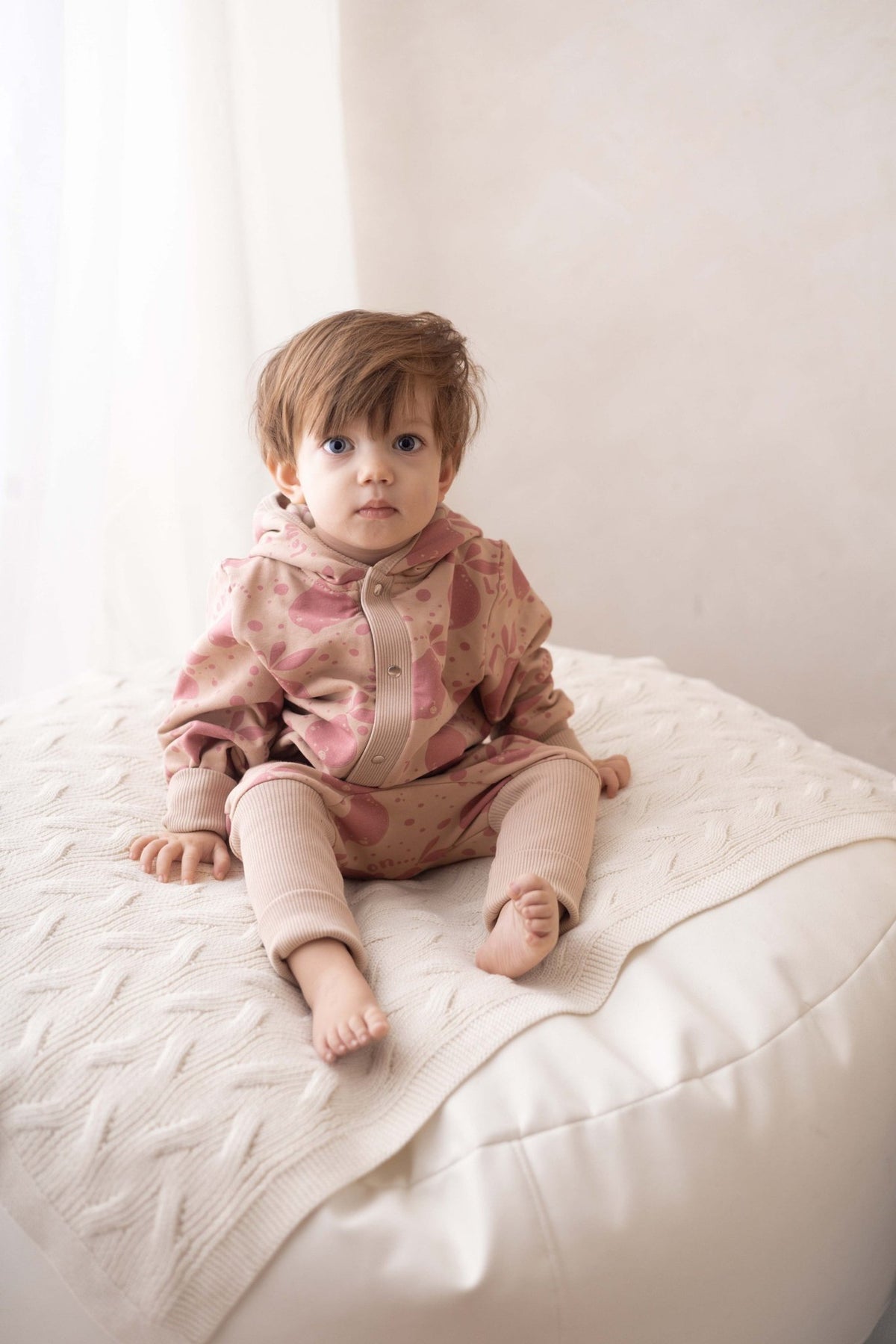 1. Baby sitting on white cushion wearing Zezuzulla beige jumpsuit with pink lemon print and hood with ears
