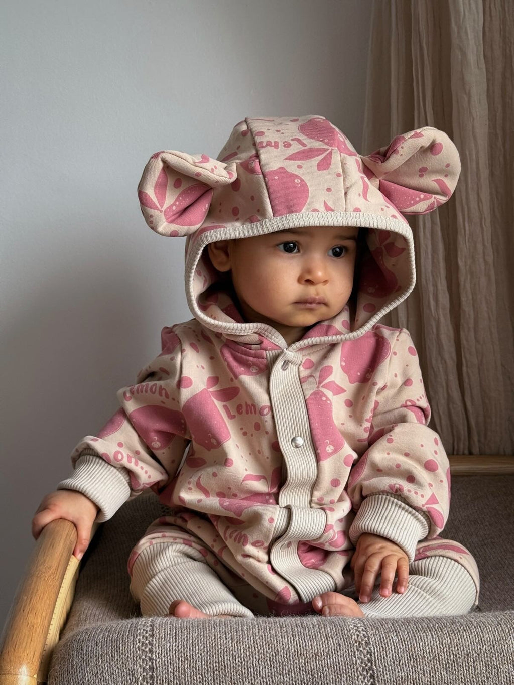 1. Baby sitting on chair wearing Zezuzulla beige jumpsuit with pink lemon print and hood with ears