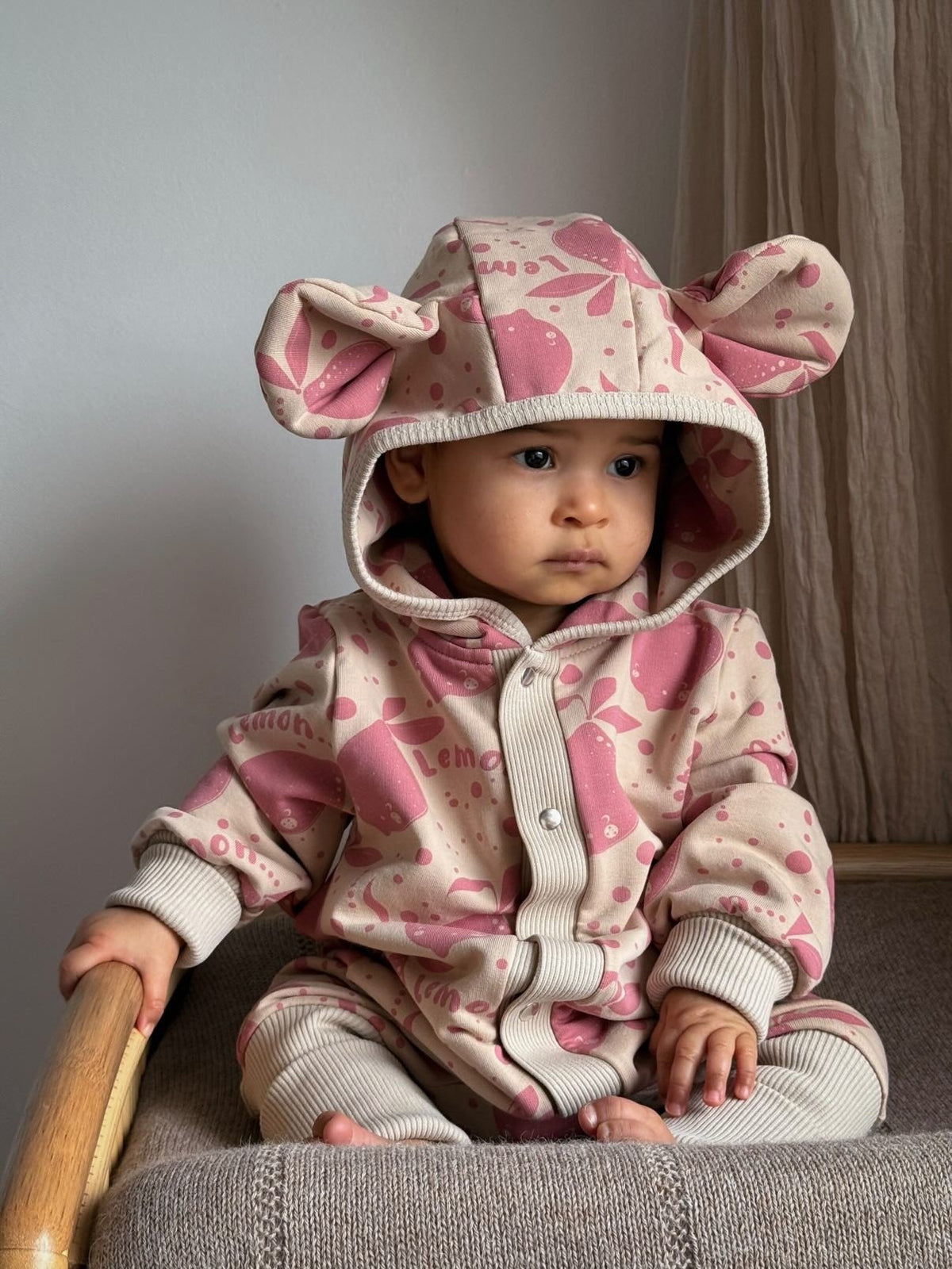 1. Baby sitting on chair wearing Zezuzulla beige jumpsuit with pink lemon print and hood with ears