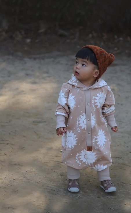 1. Baby standing on dirt path in beige sun-printed jumpsuit with hood and ears, wearing a brown beanie