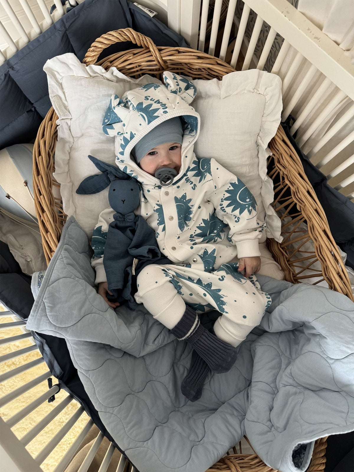 2. Infant in ecru jumpsuit with blue sun pattern and hood with ears, resting in a wicker crib with blue bedding and plush toy