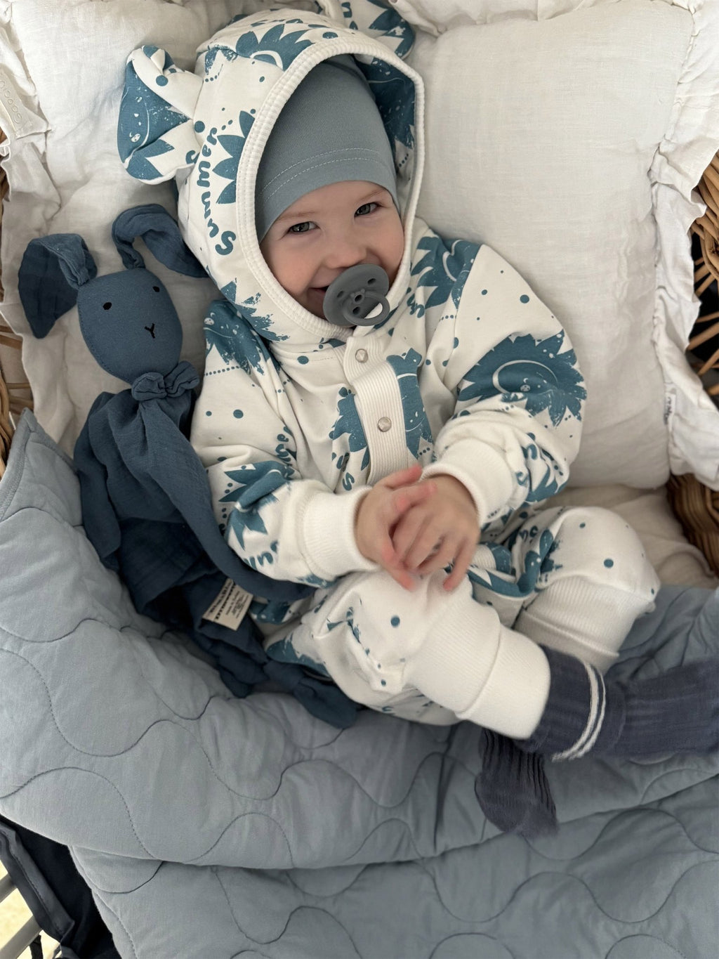 1. Baby wearing ecru jumpsuit with blue sun print and hood with ears, lying in a wicker basket with blue blanket and toy