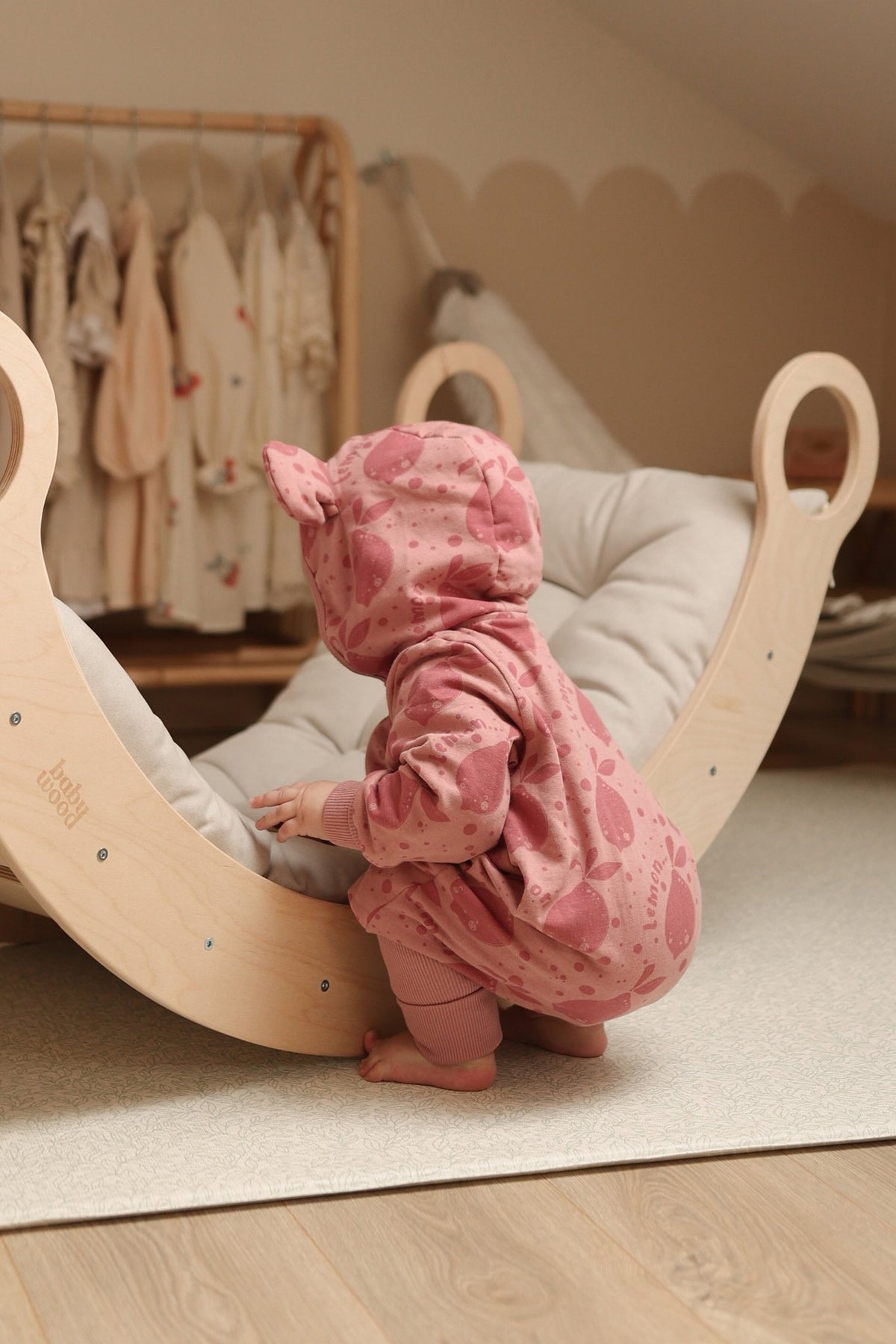 1. Baby in pink eared jumpsuit with lemon print standing on wooden rocker in nursery