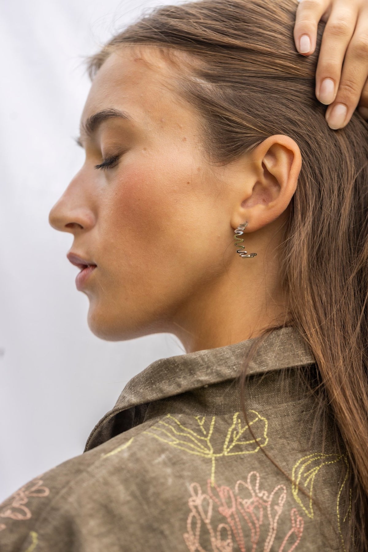 1. Woman with hand on head wearing Lentsius stainless steel wavy hoop earring, side profile, in natural light