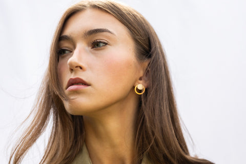1. Woman wearing Lentsius Luna Sunlit earrings with gold circular design, looking to the side in outdoor setting