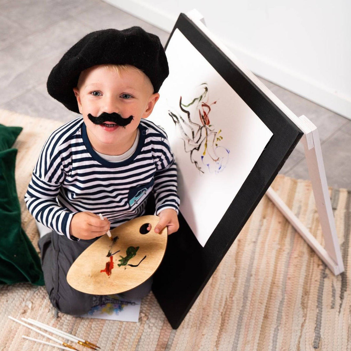 5. Child in striped shirt and beret smiling while painting on Ewart Woods black wooden chalkboard easel