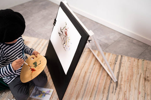 1. Child in striped shirt and beret painting on Ewart Woods black wooden chalkboard easel with art supplies on floor