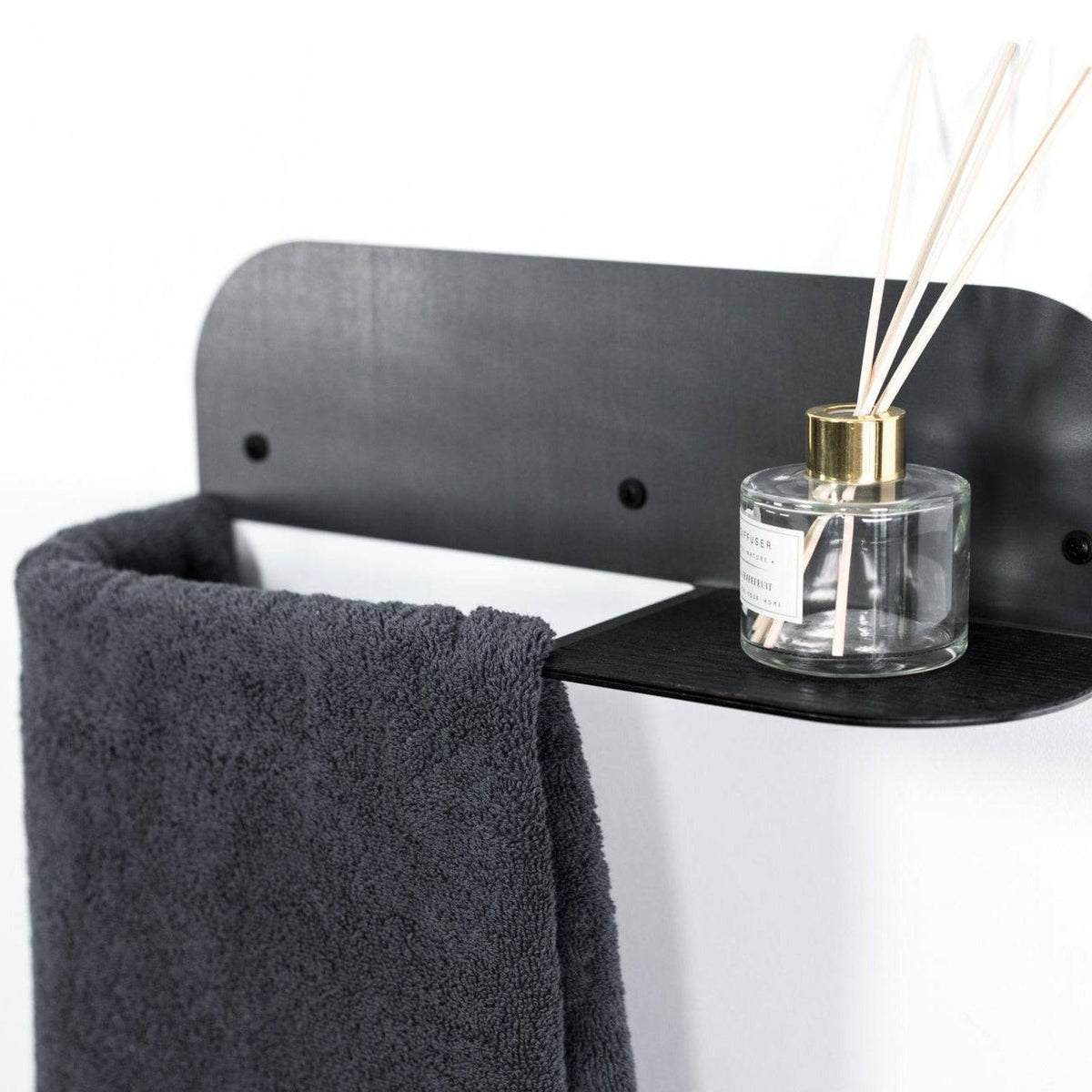 1. Wall-mounted steel and wood towel rack with black finish, holding a dark towel and a glass diffuser on a small shelf, against a white background