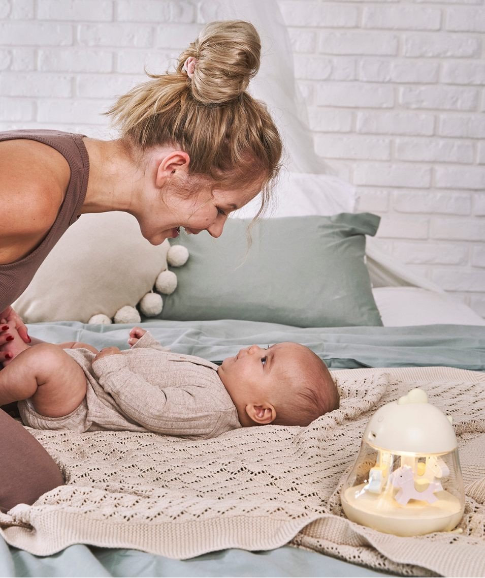 1. Woman smiling at baby on bed with yellow Rabbit & Friends lamp with carousel and music box nearby